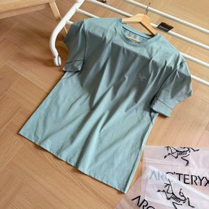 May include: A pale green short-sleeved t-shirt with a small logo on the chest. The shirt is on a wooden hanger and a white rack. Clear plastic bags with a black logo are visible.