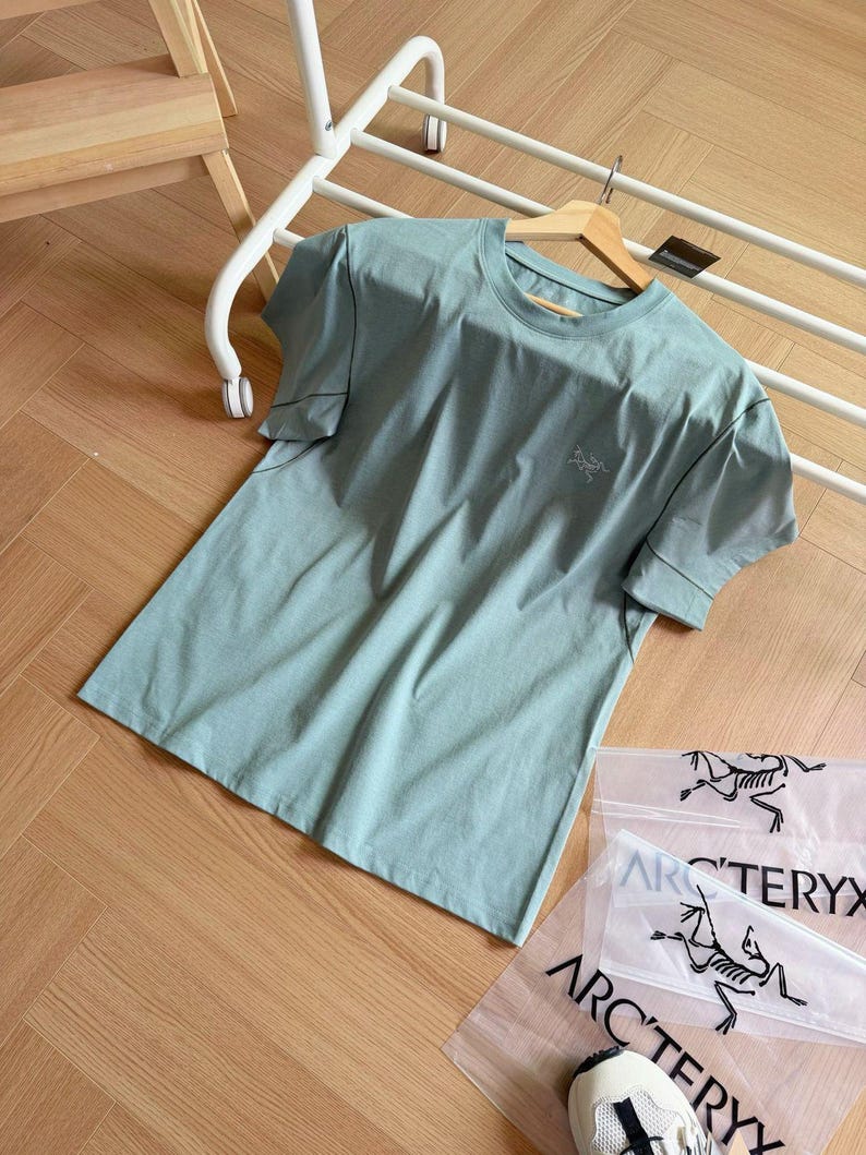 May include: A pale green short-sleeved t-shirt with a small logo on the chest. The shirt is on a wooden hanger and a white rack. Clear plastic bags with a black logo are visible.
