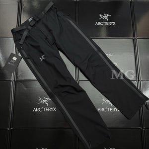May include: Black outdoor pants with a built-in belt and dark grey stripes down the sides. The pants have a zipper closure and the Arc'teryx logo is visible. The pants are displayed on a surface of black boxes.