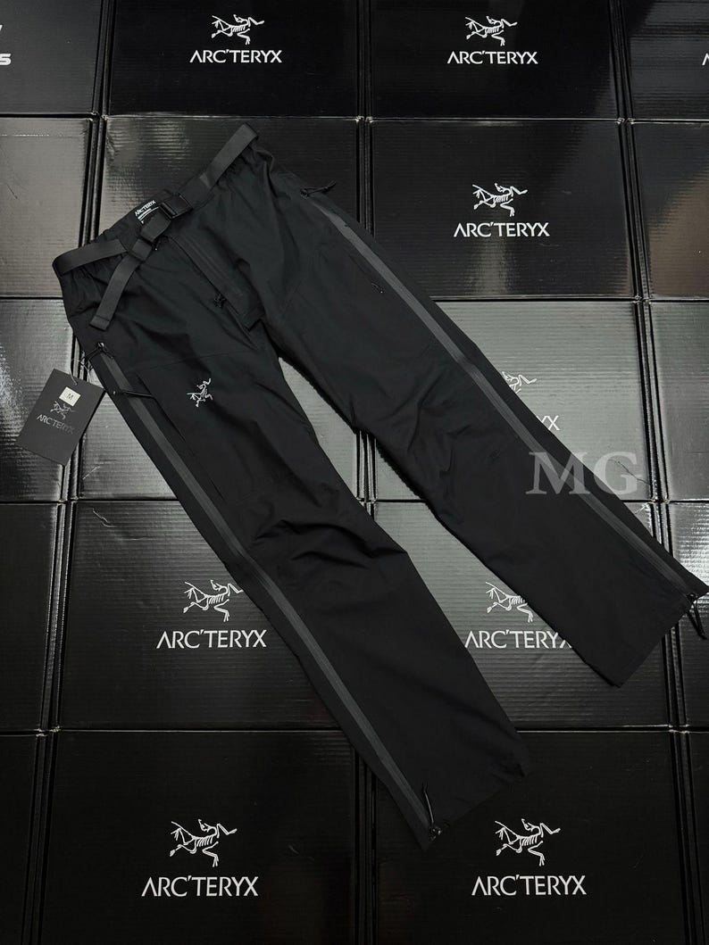May include: Black outdoor pants with a built-in belt and dark grey stripes down the sides. The pants have a zipper closure and the Arc'teryx logo is visible. The pants are displayed on a surface of black boxes.