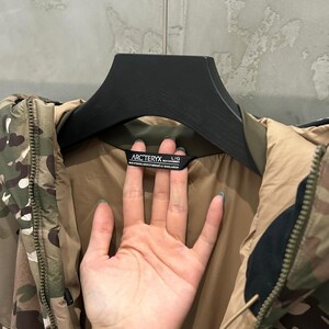 May include: Camouflage-patterned jacket with a brown interior and black accents. The jacket is hanging on a black hanger. The brand name "Arc'teryx" is visible on a tag.