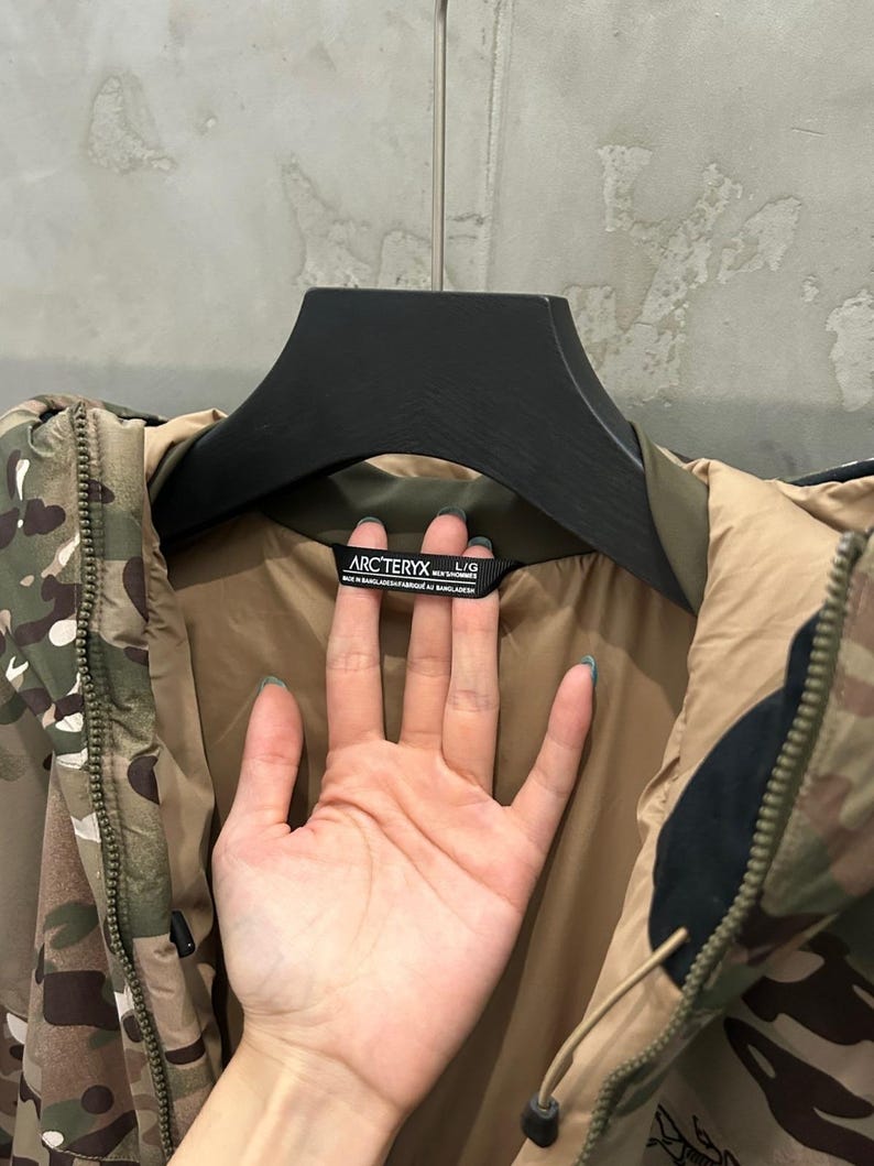 May include: Camouflage-patterned jacket with a brown interior and black accents. The jacket is hanging on a black hanger. The brand name "Arc'teryx" is visible on a tag.
