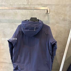 Op de afbeelding: Een marineblauwe jas met capuchon hangend aan een metalen stang. De jas heeft een minimalistisch ontwerp met een capuchon en een klein logo op de capuchon. De merknaam is zichtbaar op de capuchon. De jas lijkt gemaakt van een glad, mogelijk waterdicht materiaal.