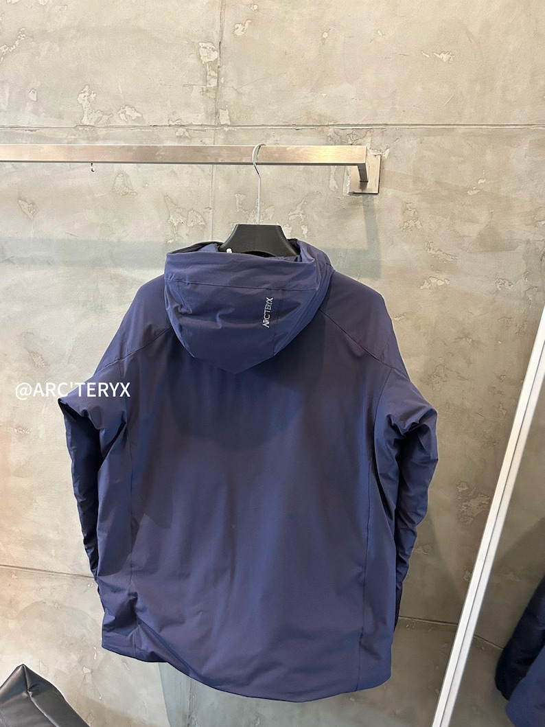 Op de afbeelding: Een marineblauwe jas met capuchon hangend aan een metalen stang. De jas heeft een minimalistisch ontwerp met een capuchon en een klein logo op de capuchon. De merknaam is zichtbaar op de capuchon. De jas lijkt gemaakt van een glad, mogelijk waterdicht materiaal.