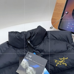 May include: Black puffer jacket with a high collar and a zip closure. The jacket features a tag with the brand name "Arc'teryx" and a yellow logo on the front. The jacket is designed for down insulation.