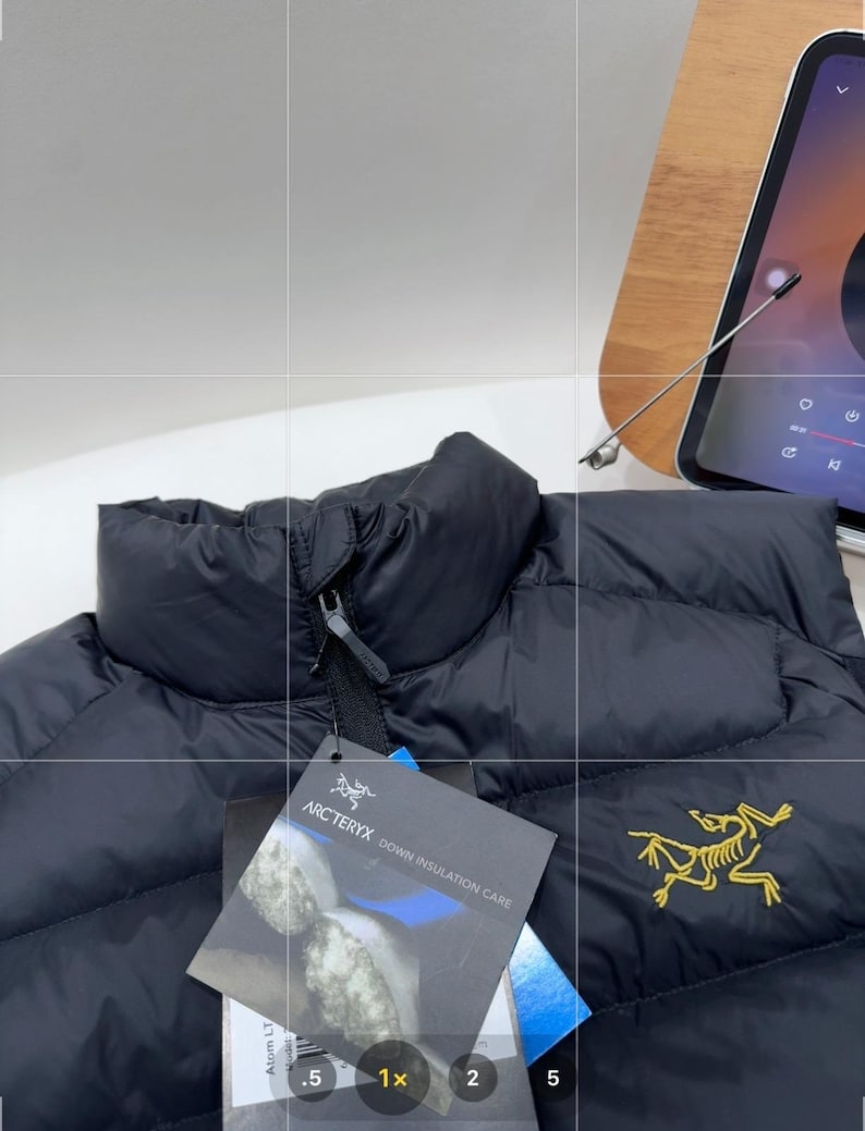 May include: Black puffer jacket with a high collar and a zip closure. The jacket features a tag with the brand name "Arc'teryx" and a yellow logo on the front. The jacket is designed for down insulation.