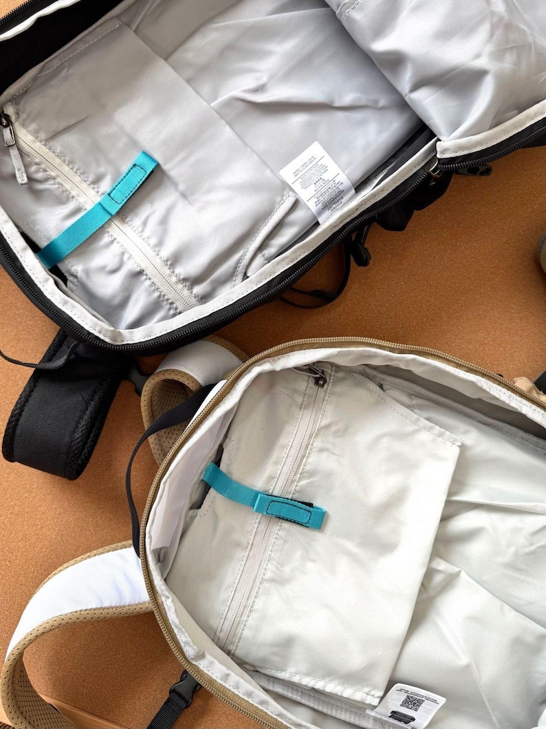 May include: Two open backpacks with interior compartments visible. One is black with a white zipper and a teal strap. The other is tan with a white strap and a teal strap. Both have a light gray interior.