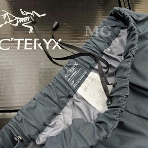 K&ouml;nnte beinhalten: Nahaufnahme einer dunkelgrauen Arc'teryx Hose. Die Hose hat einen Kordelzugbund und ein Etikett mit der Aufschrift "IN'S/HOMMES" und "S/P". Das Arc'teryx-Logo ist im Hintergrund sichtbar.