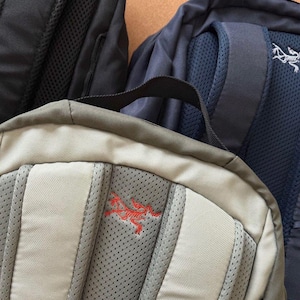 May include: Close-up of three backpacks in black, navy blue, and tan. The tan backpack has a red embroidered logo. The navy blue backpack has a white logo. All three backpacks have padded backs and shoulder straps.