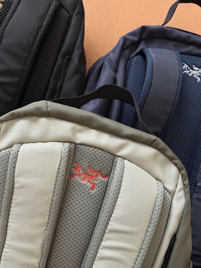 May include: Close-up of three backpacks in black, navy blue, and tan. The tan backpack has a red embroidered logo. The navy blue backpack has a white logo. All three backpacks have padded backs and shoulder straps.