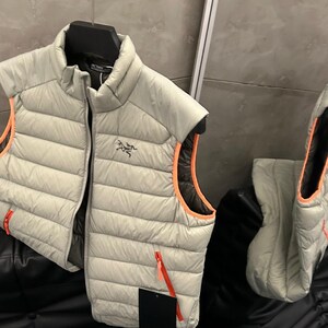 May include: A light gray puffer vest with orange trim and a red zipper. The vest is sleeveless and has a high collar. The vest is hanging on a black hanger.