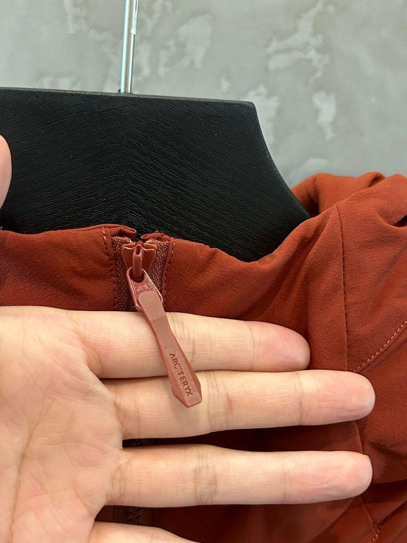 Op de afbeelding: Close-up van een roestkleurige jasrits met de merknaam "ARC'TERYX" zichtbaar op de ritstrekker. De jas hangt aan een zwarte hanger.
