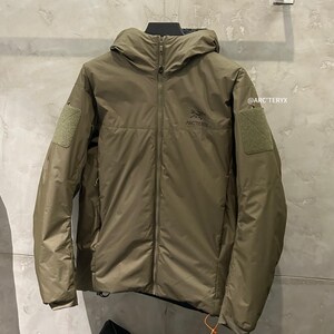May include: Olive green hooded jacket with a full-length zipper and two patch pockets on the sleeves. The jacket features the Arc'teryx logo on the chest and a small orange pull tab. The jacket is hanging on a metal bar.
