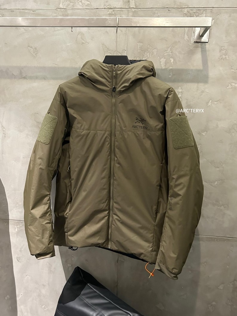 May include: Olive green hooded jacket with a full-length zipper and two patch pockets on the sleeves. The jacket features the Arc'teryx logo on the chest and a small orange pull tab. The jacket is hanging on a metal bar.