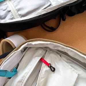 May include: Close-up of two open backpacks. One is black with a white interior and a teal strap. The other is beige with a white interior and a red zipper pull. Both have zippers and pockets.