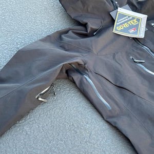 May include: Black hooded jacket with a zipper closure and multiple pockets. The jacket features a GORE-TEX tag, indicating waterproof and breathable fabric. The jacket is designed for outdoor activities and protection from the elements.