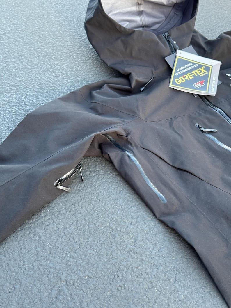 May include: Black hooded jacket with a zipper closure and multiple pockets. The jacket features a GORE-TEX tag, indicating waterproof and breathable fabric. The jacket is designed for outdoor activities and protection from the elements.