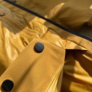 May include: Close-up of a mustard-yellow jacket with black buttons and a black zip. The jacket has a shiny, smooth texture. The buttons are embossed with the brand name.