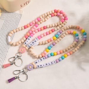 Può includere: Due cordini portachiavi con perline con i nomi "MISS SUNNY" e "MISS SHIRLEY" scritti con perline di lettere bianche. I cordini presentano perline di legno e perline di silicone colorate in tonalità pastello. Ogni cordino ha un nappa e un ciondolo a forma di fiore.