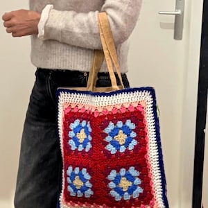 May include: A handmade tote bag with a crocheted floral design. The bag features a red center with four blue and yellow flower motifs, framed by a white, pink, and blue border. It has tan handles.