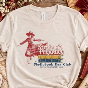 May include: A cream-coloured short-sleeve t-shirt featuring a red graphic of a person in a dress and hat running over a stack of books. The text reads Stride & Prejudice, Audiobook Run Club, and Run Til The Chapter's Done in red and blue ink.