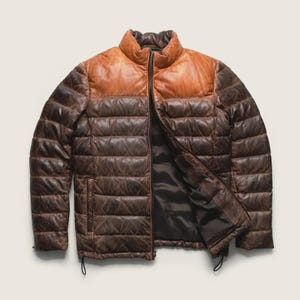 May include: A two-tone puffer jacket featuring a smooth tan leather upper section and a quilted dark brown leather lower body. The jacket has a stand-up collar, a full-length front zip, and a dark brown interior lining with visible stitching.
