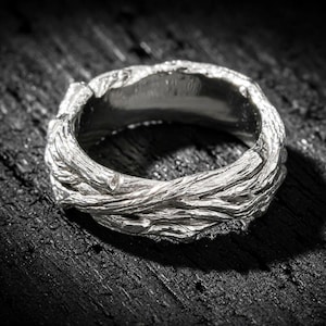 Könnte beinhalten: Ein silberner Ring mit einem strukturierten, organischen Design, das verschlungenen Ästen ähnelt. Die Oberfläche des Rings hat ein raues, rindenartiges Aussehen mit komplizierten Details und einer natürlichen Ästhetik. Der Ring ist vor einem dunklen Hintergrund platziert.