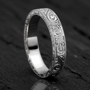 Könnte beinhalten: Ein silberner Ring mit einem aufwendigen Blumen- und Scrollmuster. Die Oberfläche des Rings ist mit detaillierten Designs graviert, wodurch ein strukturiertes Aussehen entsteht. Der Ring wird auf einer dunklen Oberfläche präsentiert, was seine reflektierenden Eigenschaften hervorhebt.