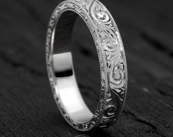May include: A silver ring with an intricate floral and scroll pattern. The ring's surface is engraved with detailed designs, creating a textured appearance. The ring is displayed on a dark surface, highlighting its reflective qualities.