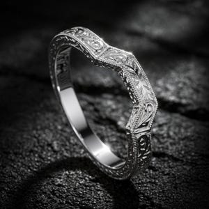 Könnte beinhalten: Ein silberfarbener Ring in Chevron-Form mit aufwendigen Gravuren von Ranken und Blattmustern auf dem äußeren Band. Die Metalloberfläche hat ein poliertes Finish mit Milgrain-Details an den Kanten, was für eine strukturierte Optik sorgt.