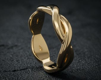 May include: A gold-colored ring with a braided design. The ring is made of a shiny, reflective metal and sits on a dark, textured surface. The ring's design features interwoven strands, creating a continuous loop.