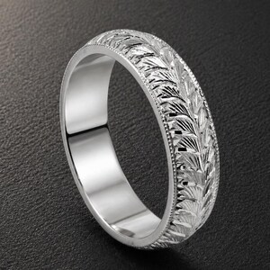 May include: A silver ring with an intricate leaf pattern engraved around the band. The ring has a polished, reflective surface and a detailed design. The ring is shown on a dark background.