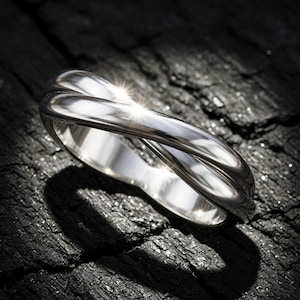 May include: A shiny silver ring with a unique intertwined design. The ring is set against a dark, textured background, with light reflecting off its polished surface. The ring's design features two bands that cross over each other.