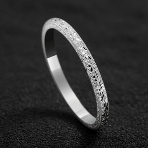 Könnte beinhalten: Ein silberner Bandring mit einem komplizierten Blumenmuster. Der Ring ist dünn und zart, mit einer polierten Oberfläche. Das Design zeigt detaillierte Blätter und Ranken, die eine Vintage-Ästhetik erzeugen. Der Ring wird vor einem dunklen Hintergrund gezeigt.