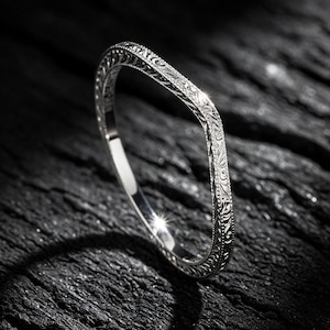 Könnte beinhalten: Ein silberner Ring mit einem aufwendigen, gravierten Blumenmuster. Der Ring ist vor einem dunklen, strukturierten Hintergrund platziert, der seine polierte Oberfläche und sein detailliertes Design hervorhebt. Die Form des Rings ist leicht gebogen.