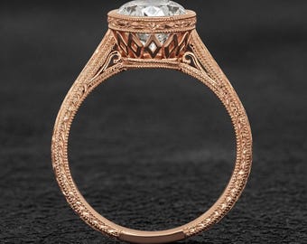 May include: Rose gold engagement ring with a large, round, clear gemstone. The ring features intricate filigree detailing around the gemstone and along the band. The band has a delicate, engraved pattern. The ring is set against a dark background.