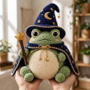 Puede incluir: Un peluche de rana tejido a ganchillo de color verde, con un sombrero de mago azul oscuro y una capa a juego, ambos decorados con estrellas doradas bordadas y una luna creciente. La rana sostiene una varita marrón con una estrella.