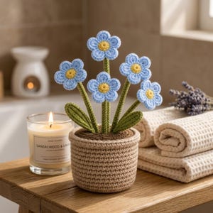 Puede incluir: Un arreglo floral de ganchillo en una maceta tejida. Las flores son de color azul claro con centros amarillos y tallos y hojas verdes. La maceta es de color marrón claro. Una vela encendida y toallas enrolladas están en el fondo.