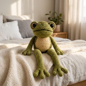 May include: A crocheted frog plush toy featuring a textured olive green body with a contrasting cream-colored belly, large black plastic eyes, and long limbs with four-toed feet, crafted with visible yarn stitches throughout the entire figure.