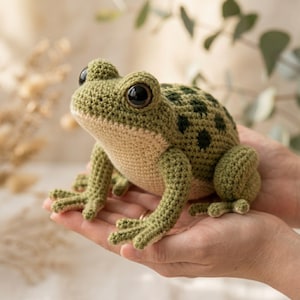 May include: A handmade crochet frog in shades of green and tan. The frog has large, dark eyes and is detailed with black spots on its back. The amphibian is held in open hands, showcasing the intricate stitch work and soft texture of the yarn.
