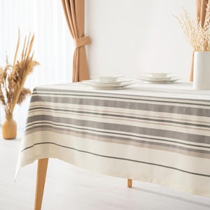 May include: A rectangular tablecloth with horizontal stripes in shades of gray and beige covers a wooden table. White plates are stacked on the table. A vase with dried plants is on the table. The room has beige curtains.