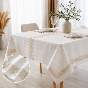 Puede incluir: Un mantel rectangular de color blanco roto con un borde bordado decorativo en tono bronceado que presenta un intrincado patrón de nudos tejidos. La tela tiene una apariencia texturizada y el diseño se repite en los bordes y como camino central.