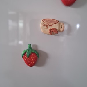 Puede incluir: Imanes de nevera pintados a mano con forma de corazón rojo, fresa y taza de café. La fresa es roja con un tallo verde. La taza de café es beige con la palabra "coffee".