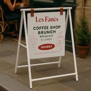Könnte beinhalten: Ein weißes A-Rahmen-Schild für ein Café mit dem Namen "Les Fanés" in Rot. Das Schild wirbt für Kaffee, Brunch, Frühstück und Mittagessen, mit dem Wort "OUVERT" in Rot. Das Schild wird von braunen Lederriemen gehalten.