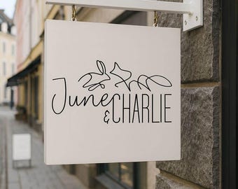 Puede incluir: Un letrero comercial cuadrado, blanquecino, con el texto "June & Charlie" en escritura negra y un dibujo de dos conejos. El letrero está suspendido de un soporte metálico blanco.