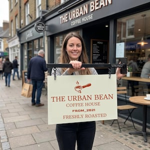 May include: A rectangular off-white sign with a black metal mounting bracket and chains. The sign features a brown graphic of a portafilter and the text: THE URBAN BEAN COFFEE HOUSE FROM_2021 FRESHLY ROASTED. The sign is held by a person.