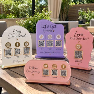 May include: Four acrylic social media signs in white, purple, and pink with gold accents. Each sign features QR codes, social media icons, and text including "Stay Connected," "Let's Get Social," "Love Our Service?," and "Follow Our Journey."