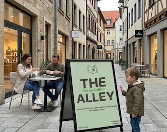 Puede incluir: Un letrero de acera negro en forma de A con una cara de color verde claro que presenta el texto THE BERLIN EDITION ALLEY, A curated selection of locally crafted goods and unique imas, Since 2018, y Ponnory Geeds, en una calle peatonal.