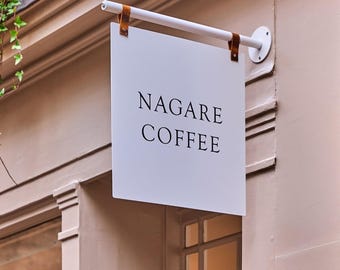 Puede incluir: Un letrero cuadrado blanco con el texto negro NAGARE COFFEE impreso en una fuente serif. El letrero cuelga de una barra metálica horizontal blanca sujeta a la pared por dos correas de cuero marrón, situado sobre el marco de una ventana.