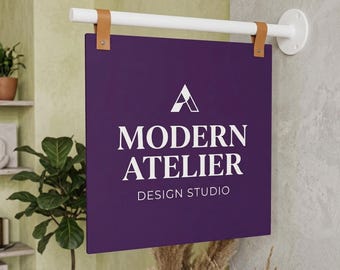 Puede incluir: Un letrero cuadrado de pared en color morado con el texto blanco MODERN ATELIER DESIGN STUDIO y un logotipo geométrico. El letrero cuelga de una barra horizontal blanca mediante dos correas de cuero beige sujetas a un soporte de pared circular.